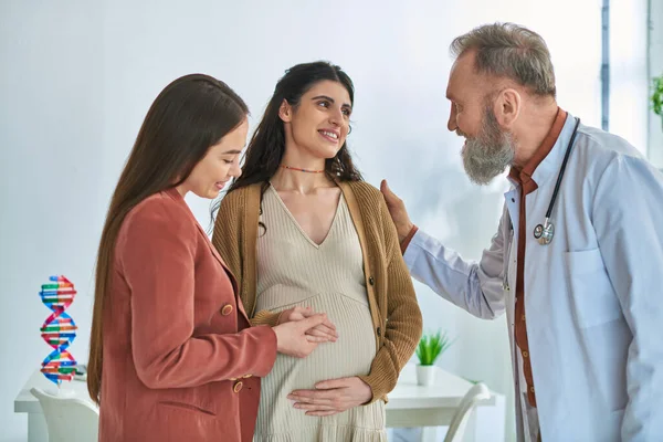 Elleri hamile karnında gülümseyen genç lezbiyen çift doktorlarına bakıyor.