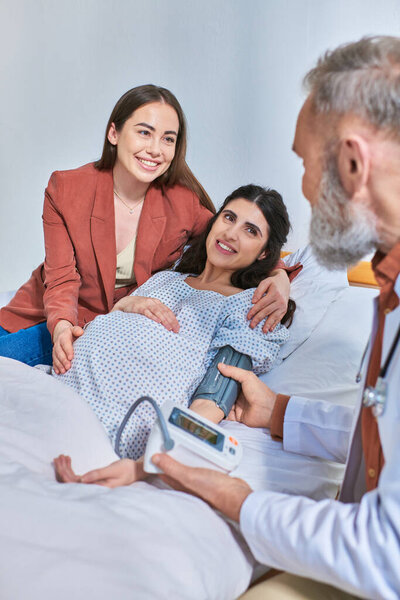cheerful lesbian couple hugging and looking at doctor that measuring pressure, ivf concept