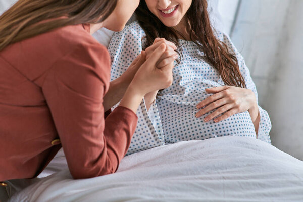 cropped view of smiling lesbian couple holding hands with one hand on pregnant belly, ivf concept