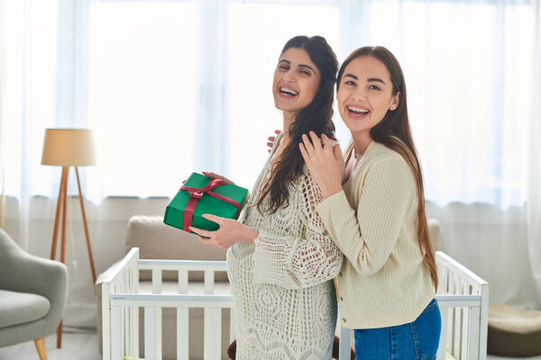 cheerful lesbian couple standing next to crib with gift in hands and smiling at camera, ivf concept