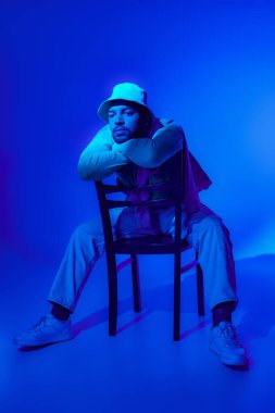 stylish african american in casual outfit sitting on chair surrounded by blue light, fashion concept clipart