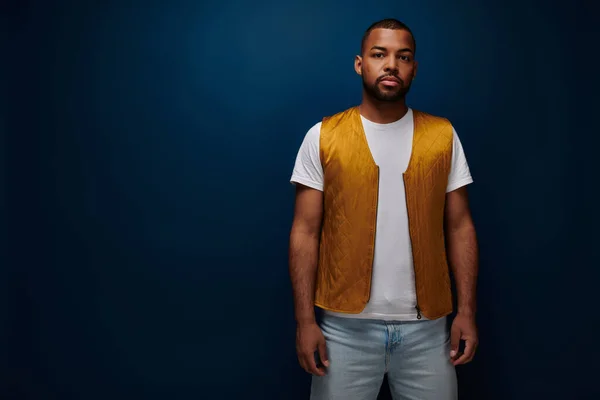 stock image handsome bearded man in yellow trendy vest standing still on dark blue backdrop, fashion concept