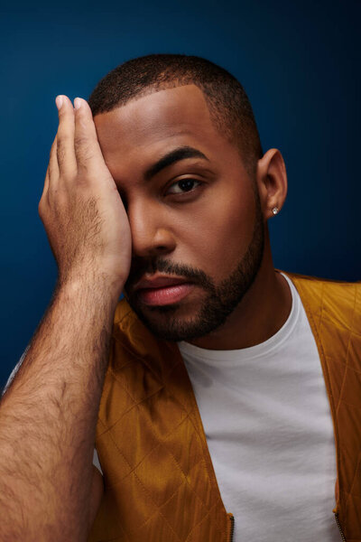 portrait of young man on dark blue backdrop with his palm on face looking at camera, fashion concept