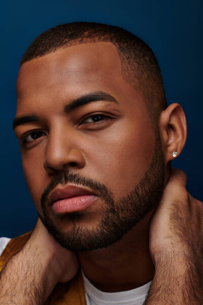 handsome man with diamond earring and beard looking at camera with hands on neck, fashion concept