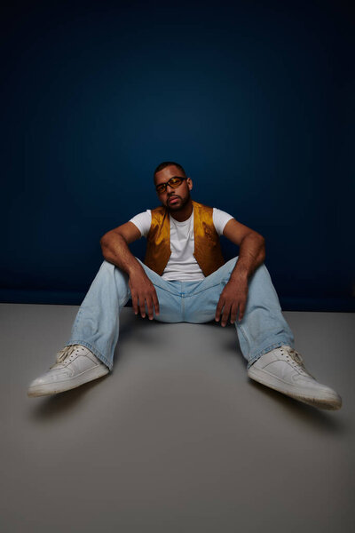 relaxed male model in yellow vest and jeans posing on floor with arms on knees, fashion concept