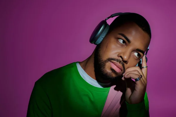 thoughtful stylish man with rings and headphones looking up on purple backdrop, fashion concept
