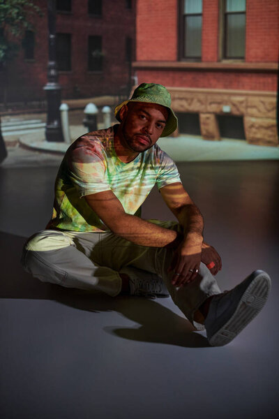 young african american man in trendy outfit sitting on floor in projector lights, fashion concept