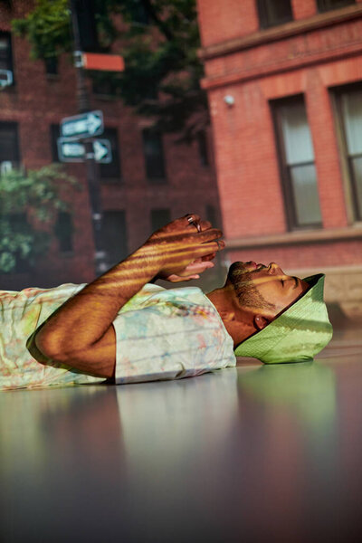 стильный африканский американец в уличной одежде, лежащий на полу в проекторе огней, концепция моды