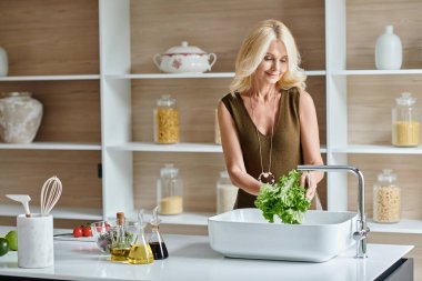 carefree middle aged vegetarian woman with blonde hair washing fresh lettuce while preparing salad clipart