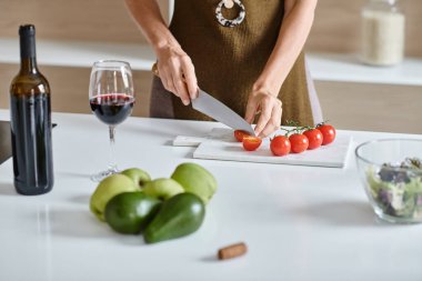 Kırpılmış kadın kırmızı şarap, avokado ve yeşil elmaların yanında taze vişneli domates kesiyor.