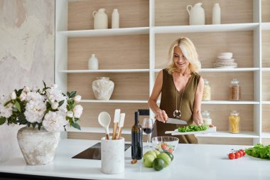 Modern mutfakta taze malzemelerden salata yapan orta yaşlı çekici bir kadın.