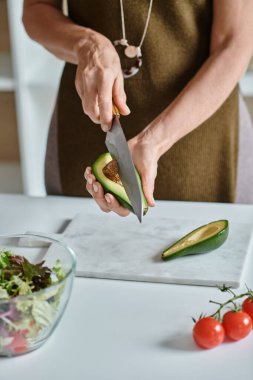 cropped woman cutting fresh avocado half near cherry tomatoes and lettuce in bowl, home cooking clipart