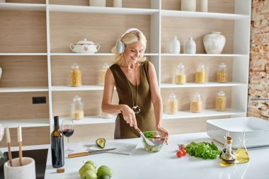 happy middle aged woman in wireless headphones listening music and making salad in kitchen clipart