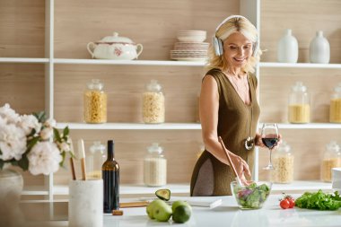 happy middle aged woman in headphones holding glass of wine and standing near fresh salad in bowl clipart