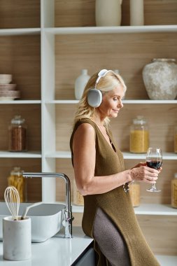 Modern mutfakta elinde bir bardak kırmızı şarap tutan orta yaşlı, kablosuz kulaklıklı bir kadın.