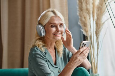 Kablosuz kulaklıklı neşeli orta yaşlı kadın müzik dinliyor ve evde akıllı telefon kullanıyor.