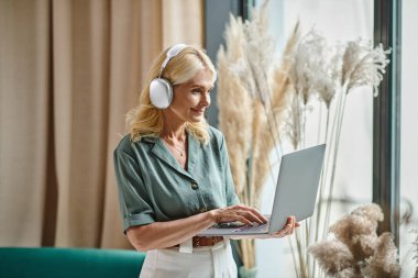 Kablosuz kulaklıklı neşeli orta yaşlı kadın müzik dinliyor ve evde dizüstü bilgisayar kullanıyor.