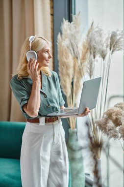 Kablosuz kulaklık takan, müzik dinleyen ve evde dizüstü bilgisayar kullanan orta yaşlı pozitif bir kadın.