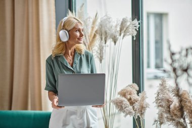 Orta yaşlı, kablosuz kulaklıklı, müzik dinleyen ve dizüstü bilgisayarını evde tutan mutlu bir kadın.