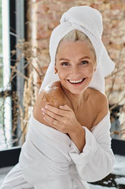 cheerful middle aged woman with white towel on head and bathrobe applying body scrub, vertical clipart