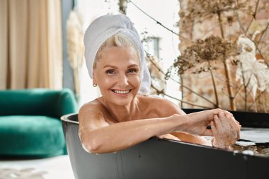 Kafasında beyaz havluyla orta yaşlı mutlu bir kadın modern bir dairede banyo yapıyor, rahatlıyor.