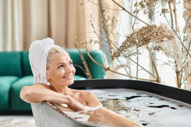 Kafasında beyaz havluyla orta yaşlı neşeli bir kadın modern bir dairede banyo yapıyor, güzellik rutini.