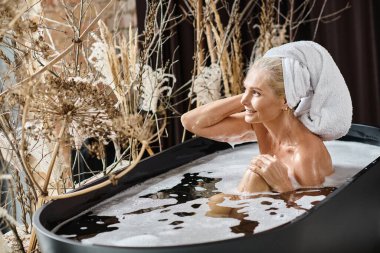 relaxed middle aged woman with white towel on head taking bath in modern apartment, beauty routine clipart