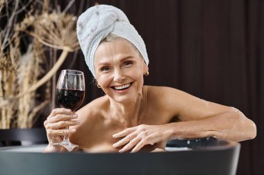 Kafasında havlu olan neşeli orta yaşlı kadın evde banyo yaparken elinde bir bardak kırmızı şarap tutuyor.