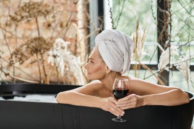 Kafasında beyaz havluyla rahat ve hayalperest bir kadın elinde bir bardak kırmızı şarapla banyo yapıyor.