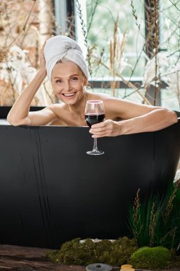 radiant middle aged woman with white towel on head holding glass of red wine and taking bath clipart