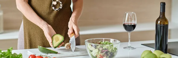 stock image cropped banner, woman cutting ripe avocado near fresh ingredients and red wine, home cooking