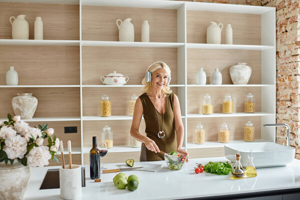 cheerful middle aged woman in wireless headphones listening music and making salad in kitchen