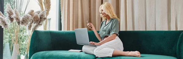 快乐中年妇女手握智能手机 坐在沙发上使用笔记本电脑的横幅 — 图库照片