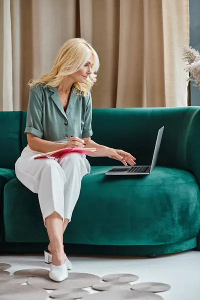 stock image blonde middle aged woman using laptop and taking notes while sitting on sofa in modern living room