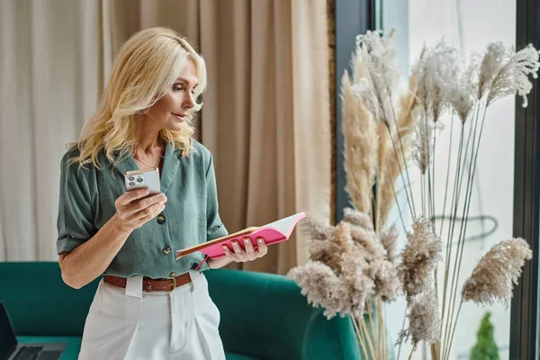 stock image blonde middle aged woman looking at notes in notebook and holding smartphone while standing at home