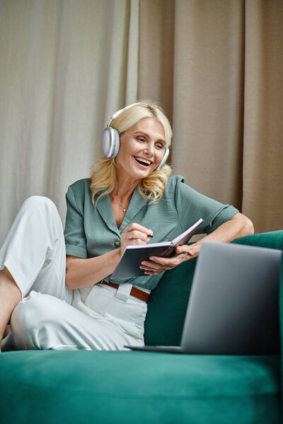 excited middle aged woman in wireless headphones sitting on sofa near laptop and writing in notebook