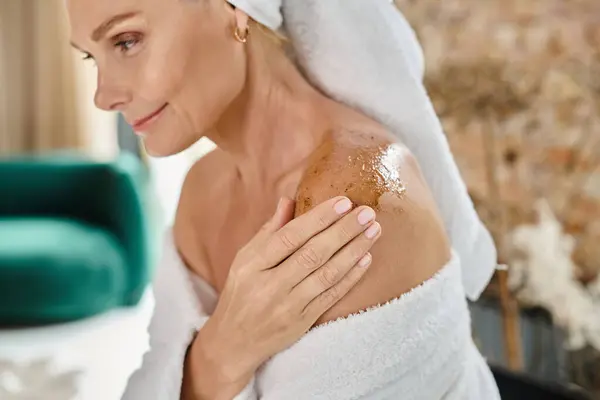 beautiful middle aged woman with white towel on head and bathrobe using body scrub on shoulder