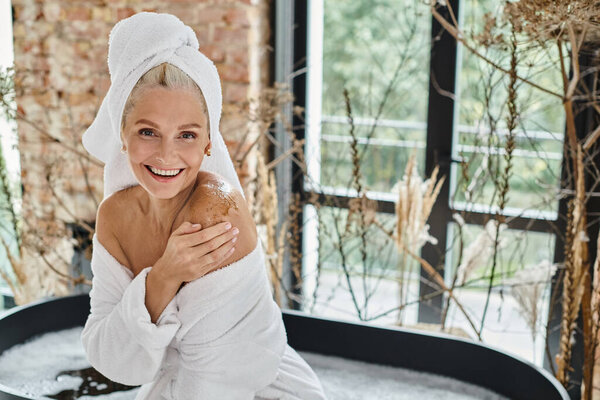 cheerful middle aged woman with white towel on head and bathrobe applying body scrub near bathtub