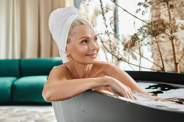 pleased middle aged woman with white towel on head taking bath in modern apartment, relaxation