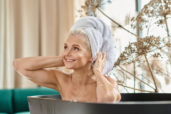 cheerful middle aged woman with white towel on head taking bath in modern apartment, home spa