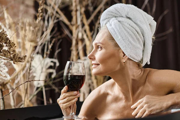 Mutlu orta yaşlı kadın kafasında havluyla elinde bir bardak kırmızı şarapla evde banyo yapıyor.