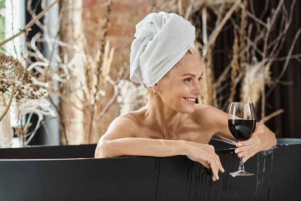 calm and happy middle aged woman with towel on head holding glass of red wine while taking bath