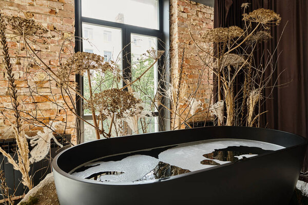 water inside of black bathtub in modern apartment with windows and decorative plants