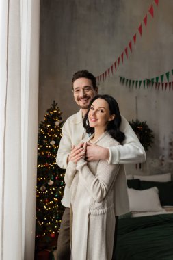 positive man hugging wife in cozy home wear near blurred decorated Christmas tree, winter holidays clipart