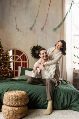 Mutlu evli çift, modern apartmandaki süslü Noel ağacının yanında sarılıp yatıyorlar.