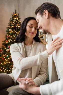 happy woman with closed eyes sitting with husband near blurred Christmas tree, season of joy clipart