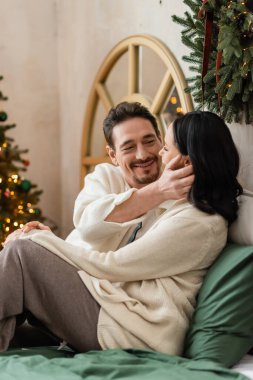Neşeli adam, karısının yüzüne nazikçe dokunuyor. Noel ağacının bulanık ışıkları yanında, sıcacık bir sabah.