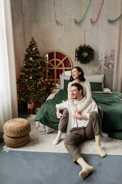 Kış tatili, mutlu kadın süslü yatak odasında Noel ağacı ve çelenkle kocasını kucaklıyor.