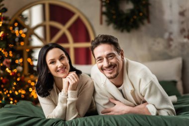 Mutlu evli bir çiftin portresi kameraya bakıyor ve Noel ağacının yanındaki yatakta uzanıyor.