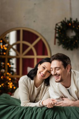 Mutlu evli bir çiftin portresi kameraya bakıyor ve Noel ağacının yanındaki yatakta uzanıyor.